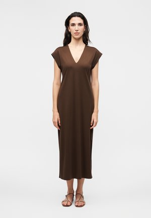 Moteris stovi, vilki be rankovių rudą midi su V formos iškirpte suknelę ir rudus sandalėlius, prieš šviesų vientisą foną.