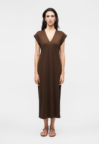 Woman standing, wearing a sleeveless brown V-neck midi dress and brown sandals, against a plain light background.