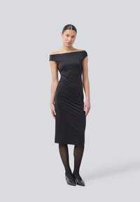 Svart off-the-shoulder kjole laget av glatt stoff, tettsittende design, kne-lengde, med subtil rynking på siden, båret med svarte strømpebukser og hæler.
