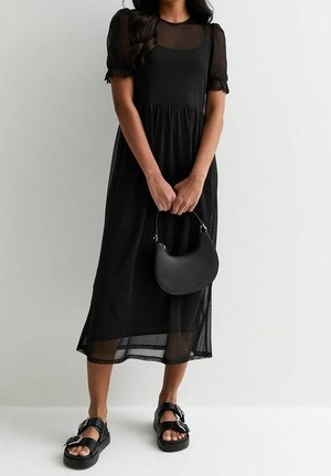 Mujer con un vestido midi negro con mangas cortas y dobladillo transparentes, sosteniendo un bolso negro y llevando sandalias negras con hebilla.