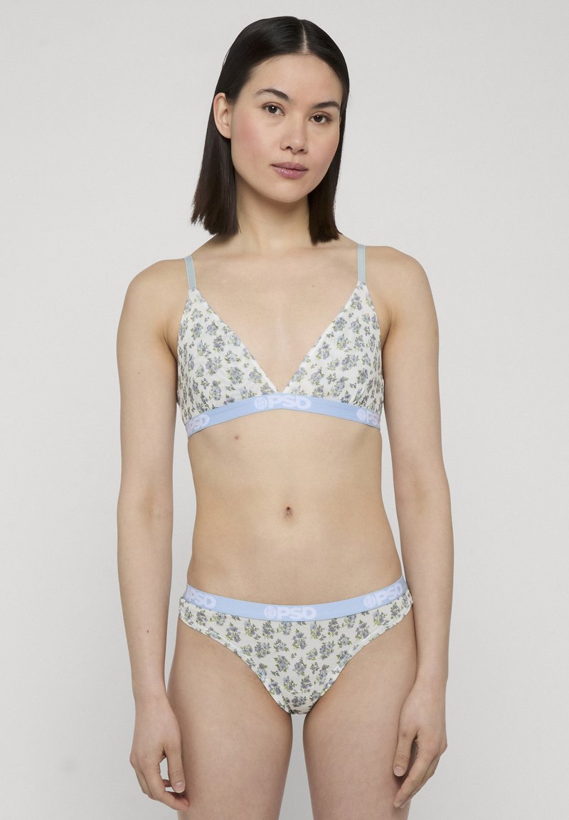 Jeune femme aux cheveux lisses et foncés, portant un soutien-gorge blanc à fleurs et un slip assorti avec des bandes élastiques bleu clair, debout devant un fond uni.