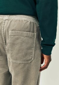 Helle graue Cordhosen mit vertikalen Streifen, ausgestattet mit einer Gesäßtasche und einem elastischen Bund. Der Stoff wirkt weich und strukturiert.