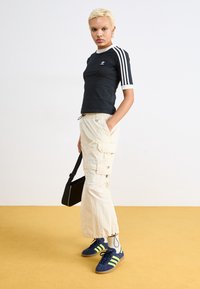 Črna športna majica z belimi obrobami in tremi belimi črtami na rokavih, bež cargo hlače z stranskimi žepi, mornarsko modri čevlji z rumenimi črtami.