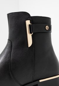 Bottine en cuir noir avec une finition texturée, un accent doré sur le côté et un design épuré et structuré.