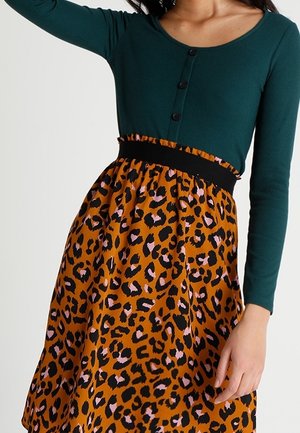 Vrouw die een teal kleurige top met lange mouwen draagt en een hoog getailleerde oranje rok met een zwart-roze luipaardprint.