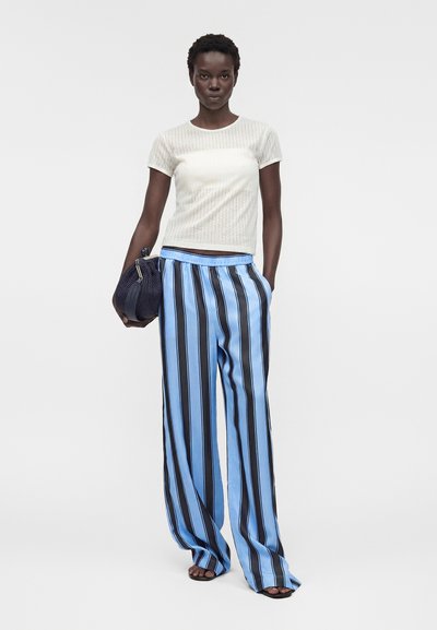 Γυναίκα που φοράει λευκό τοπ με κοντά μανίκια και φαρδιά μπλε παντελόνια με μαύρες κάθετες ρίγες, κρατώντας μια μπλε υφασμάτινη τσάντα φάκελο.