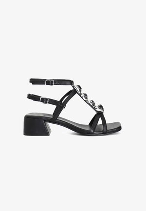 Sandalo in pelle nera con tacco a blocco, dotato di cinturini alla caviglia regolabili e decorazioni in strass sul dorso del piede, con un design minimalista.