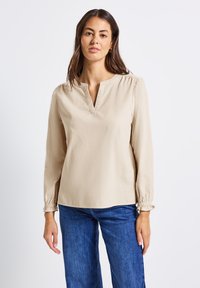 Frau mit langen braunen Haaren, die ein beiges Longsleeve-Oberteil mit ausgeschnittenem Ausschnitt und blaue Jeans trägt, vor einem schlichten weißen Hintergrund.