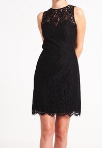 Robe sans manches en dentelle noire avec un motif floral, taille cintrée et ourlet festonné, portée par une personne se tenant avec les mains derrière le dos.