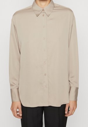 Person wearing a beige long-sleeve button-up shirt with a pointed collar and black pants, standing against a plain background.