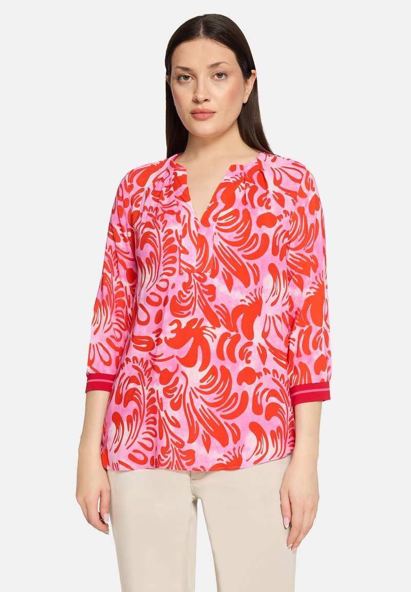 Blouse à motif floral en rose et rouge, confectionnée dans un tissu léger. Elle présente un décolleté en V et des manches trois-quarts avec des accents rouges.