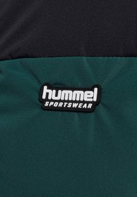 Hummel Sportswear logo patch op een zwarte en donkergroene stoffen achtergrond, met een kledingmerkenlabel.
