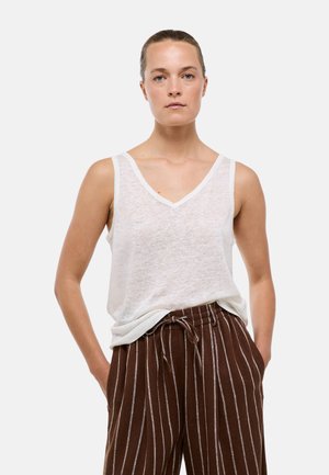 Jeune femme aux cheveux attachés en arrière, portant un haut blanc sans manches à col en V et un pantalon ample marron à rayures, les mains dans les poches.
