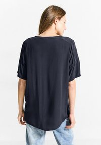 Tmavě šedé oversized tričko s kulatým výstřihem a krátkým rukávem. Tkanina je hladká s jemným leskem a má zakřivený lem.