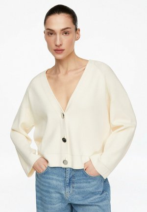 Kobieta o ciemnych włosach, w kremowym swetrze na guziki i niebieskich jeansach, z rękami w kieszeniach, na jednolitym białym tle.