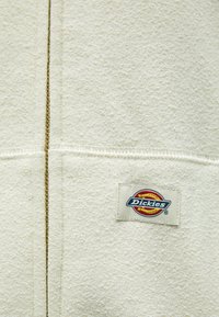 Fehér bolyhos dzseki texturált anyagból, középső varrással és téglalap alakú, színes "Dickies" logó felvarrással.