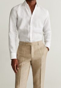 Biała lniana koszula z długim rękawem i kołnierzykiem, sparowana z beżowymi spodniami o dopasowanym kroju z zapięciem na guzik i plisami z przodu.