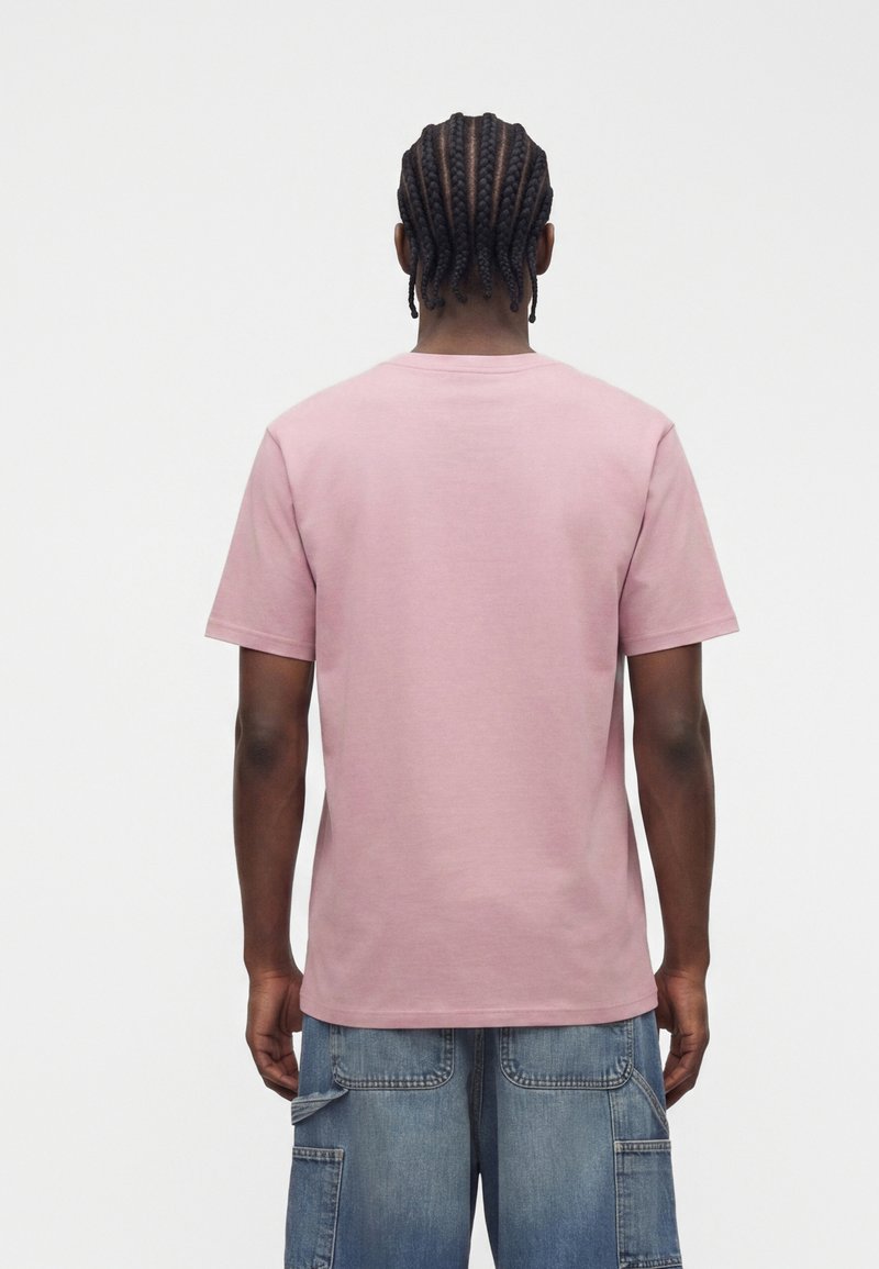 Man with braided hair wearing a plain pink t-shirt and blue denim cargo shorts, standing facing away on a white background.