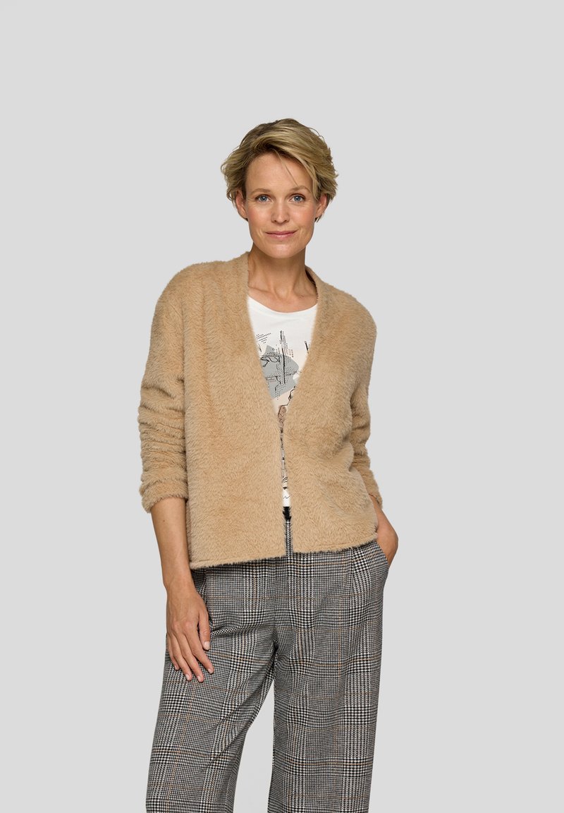 Beige flauschige Strickjacke mit entspanntem Schnitt und offenem Vorderteil. Kombiniert mit grauen, karrierten Hosen mit weiter Silhouette.