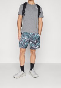 Šedé tričko, vzorované tyrkysové šortky, černé ponožky a světle šedé tenisky. Batoh na ramenou, stojí na bílém pozadí.