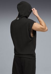 Άνδρας ρυθμίζει τη κουκούλα σε μαύρο αμάνικο φούτερ με έμβλημα τίγρης στην πλάτη, φοράει αντίστοιχο μαύρο παντελόνι πάνω σε γκρίζο φόντο.