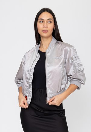 Vrouw die een glanzend zilveren bomberjack draagt over een zwarte jurk, het ritsje van het jack vasthoudend en zijwaarts kijkend.