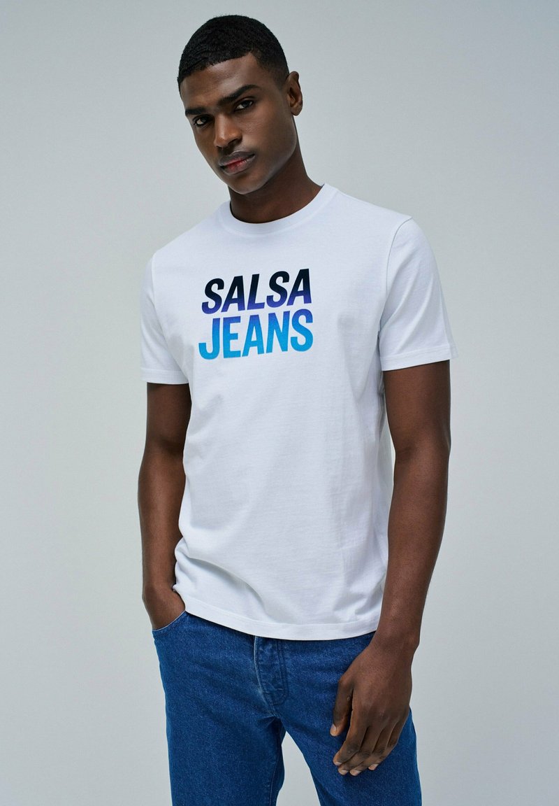 Camiseta blanca de algodón con un gráfico degradado en azul que dice "SALSA JEANS", combinado con pantalones de mezclilla azul oscuro. Tiene un cuello redondo sencillo y mangas cortas.