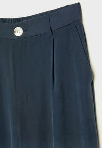 Pantalon bleu foncé en gros plan montrant la ceinture avec un bouton blanc, passants pour ceinture, poche latérale et détail du pli avant.