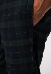 Geruite broek in donker groen en marineblauwe stof met een zachte textuur. Heeft zijzakken en een subtiele geborduurde logo.