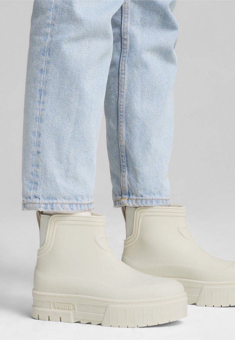 Personne portant un jean bleu clair et des bottines en caoutchouc blanc cassé avec des semelles épaisses sur un fond blanc.