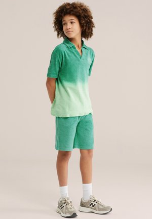Kind met krullend haar, gekleed in een groene polo met kleurverloop en bijpassende korte broek, staand met de handen achter de rug, naar de zijkant kijkend.