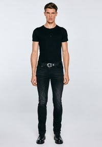 Jonge man staande, gekleed in een zwart kortärmelig henley-shirt, zwarte jeans met riem en zwarte veterlaarzen, tegen een eenvoudige lichte achtergrond.