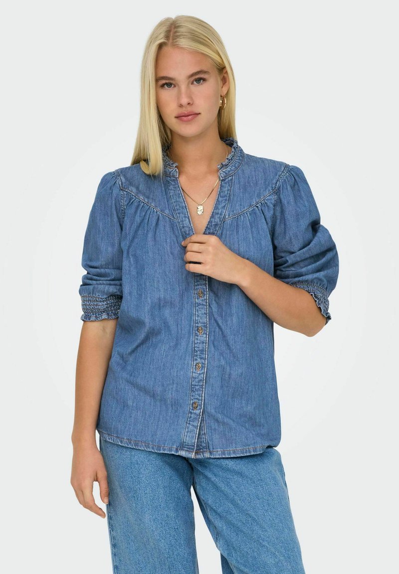 Camicia in denim blu chiaro con chiusura a bottoni, yoke plissettato e maniche a palloncino. Presenta un colletto arricciato e elastico ai polsi delle maniche.