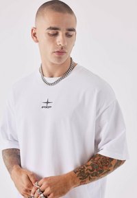 Witte katoenen T-shirt met korte mouwen en een ronde hals, met een klein zwart logo op de borst. Model draagt een dikke zilveren ketting.