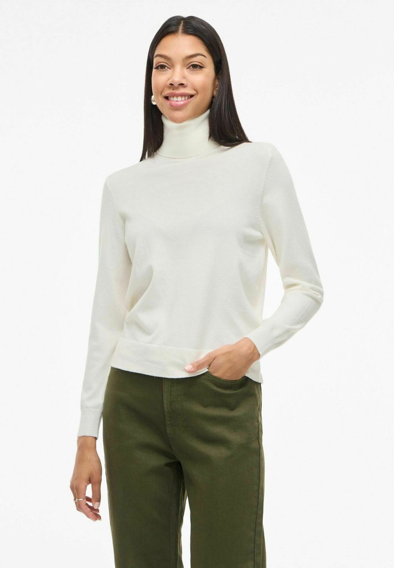 Jersey de cuello alto crema con mangas largas, textura suave y diseño ajustado, combinado con pantalones verde oliva. Estilo simple y minimalista.