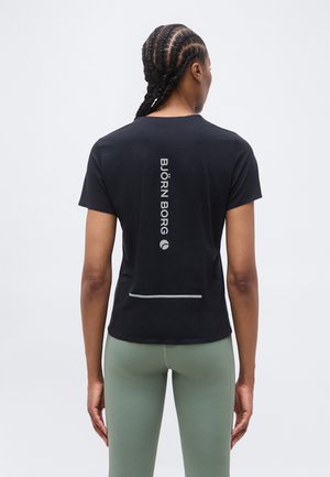 Γυναίκα με πλεγμένα μαλλιά που φοράει μαύρο μπλουζάκι Björn Borg και ανοιχτοπράσινες κολάν, γυρισμένη με την πλάτη σε απλό φόντο.