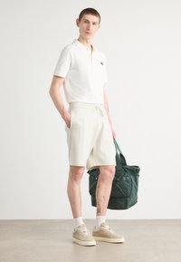 Camiseta polo blanca, pantalones cortos a rayas claros, zapatillas beige y bolsa verde. Modelo de pie con las manos en los bolsillos, mostrando un atuendo minimalista.