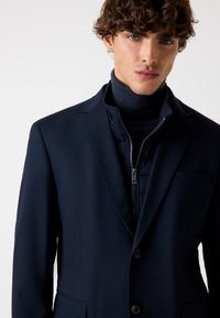 Marineblauwe blazer met notch lapel, een enkele knoopsluiting en een gladde stof. Gelaagd over een marineblauwe gewatteerde jas met rits.