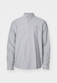 Camicia a maniche lunghe grigia chiara con bottoni e colletto a camicia, dotata di un piccolo logo rosso, bianco e blu sulla parte sinistra del petto.