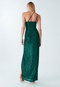 Emeraldų žalia maxi suknelė iš blizgančios, siuvinėtos blizgučių audinio. Suknelė turi plonas petnešėles, prigludusią liemenį ir šoninius plyšius judėjimui.