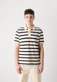Polo-shirt i kremfarge med marineblå horisontale striper. Har krage og korte ermer. Diskret logo på venstre side.