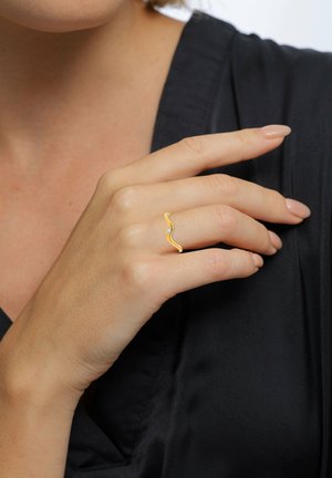 A close-up of a hand displaying a gold wavy ring with a small stone, set against a dark fabric background. The nail polish is nude.
