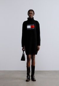 Musta neulottu poolomekko, jossa näkyvä "TOMMY JEANS" -logo punaisena, valkoisena ja sinisenä. Yhdistetty pieniin mustiin käsilaukkuun ja polvipitkiin saappaisiin.