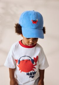 Casquette de baseball en coton bleu avec un crabe rouge brodé portant des lunettes de soleil et un chapeau. T-shirt blanc avec col rouge, arborant un motif de crabe.
