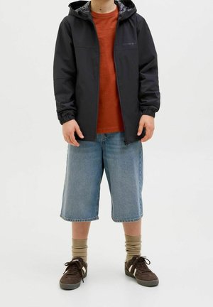 Niño con chaqueta negra con capucha, camiseta naranja, pantalones cortos de mezclilla hasta la rodilla, calcetas beige y zapatillas marrones, de pie contra un fondo blanco.