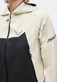 Veste beige et noire zippée avec capuche, avec un logo animal stylisé sur la poitrine et le texte « DYNA » sur la manche.