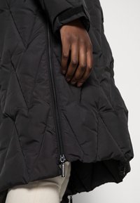 Veste matelassée noire avec motif matelassé diagonal, fermeture éclair à l'avant et poignets en velcro. Une main tient la fermeture éclair, mettant en valeur les détails du design.
