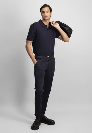 Vīrietis tērpies tumši zilā polo kreklā un biksēs, melnā jostā un apavos, ar melnu somu pār plecu, stāvot gaiši pelēkā fona priekšā.