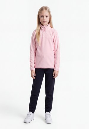 Fille blonde debout portant une veste polaire rose pâle, un pantalon noir et des baskets blanches sur fond blanc.