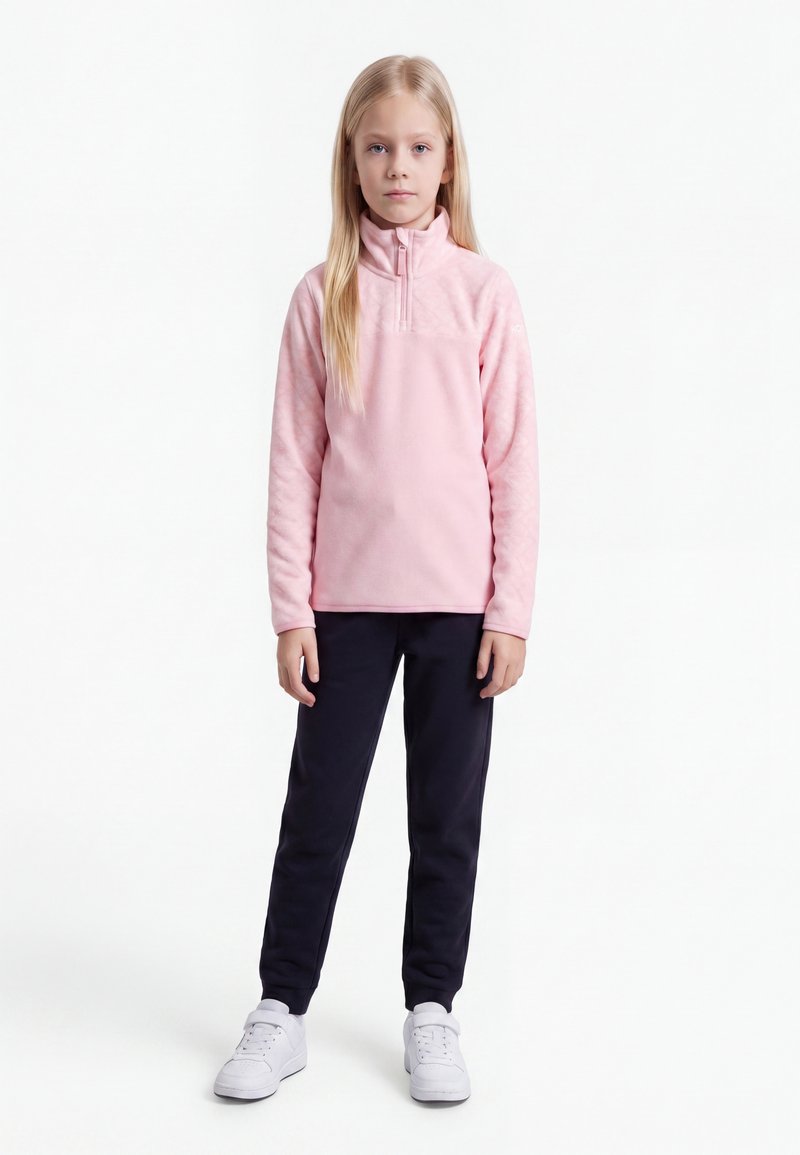 Fille blonde debout portant une veste polaire rose pâle, un pantalon noir et des baskets blanches sur fond blanc.
